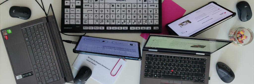 Auf einem weißen Tisch tummeln sich 2 Tablets und 2 Laptops, dazwischen 4 Mäuse, eine Tastatur mit extragroßen Tasten, farbiges Papier und ein Glas mit Süßigkeiten.