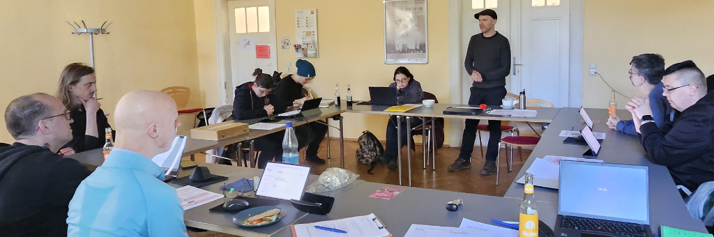 8 Personen, die an Laptops und Tablets arbeiten, sitzen an einem Konferenz-Tisch in O-Form. Eine Person mit Kappe steht und moderiert. Auf dem Tisch sind noch weitere Computer, Tagungs-Unterlagen, Snacks und Leichte-Sprache-Karten verteilt.